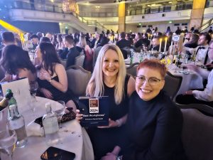 Two female colleagues sit at a table at an awards event in the evening. One of them is holding a UK's Best Workplaces trophy.